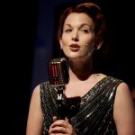 Rebecca LaChance, a white woman, sings into an old fashioned microphone while wearing a black sequined flapper dress. Her hair is pulled back in a 1940s style.